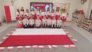 Dziewczynki i chłopcy w strojach narodowych pozują do zdjęcia na tle położonej flagi.
