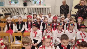 Dzieci w strojach ludowych siedzą w rzędach na drewnianych krzesełkach.