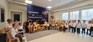 Dzieci z grup specjalnych siedzą na żółtych krzesełkach na tle granatowego materiału z napisem Pasowanie na Przedszkolaka.