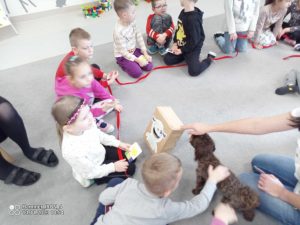 Dzieci siedzą na szarym dywanie w kole. Pomiędzy nimi chodzi brązowy pies.