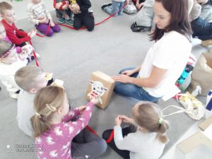 Dzieci siedzą w kole na szarym dywanie, trzymają czerwony sznurek. Przed nimi jest pudełko szare z naklejonym psem. W śród nich klęczy pani w białej bluzce.