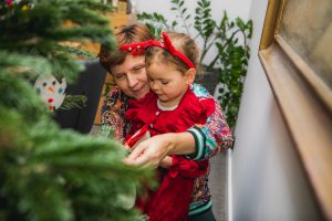 Kobieta trzymająca na kolanach dziewczynkę wieszającą ozdobę na choinkę.