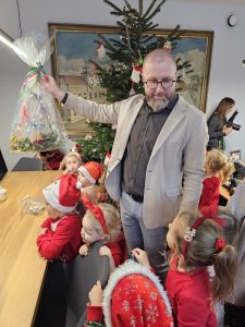 Prezydent Miasta Ostrowa Wielkopolskiego oraz dzieci ubrane w mikołajkowe stroje stojący w biurze. Pan Prezydent trzyma w ręku upominek. W tle Pani z aparatem fotograficznym.