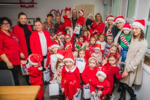 Grupa dzieci ubranych na czerwono w mikołajkowych czapkach wraz z dziewięcioma paniami oraz jednym panem.