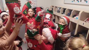 Dzieci ubrane na czerwono w czapkach Mikołaja stoją w szatni między półkami i pokazują skarpety z prezentami.