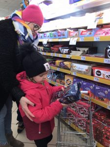 Dziewczynka z pomocą nauczyciela wybiera produkty z półki i wkłada je do koszyka.