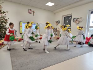 Dziewczynki ubrane na biało, na głowach kwiatki i liście na rękach tańczą na szarym dywanie.