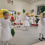 Dziewczynki ubrane na biało, na głowach kwiatki i liście na rękach tańczą na szarym dywanie.