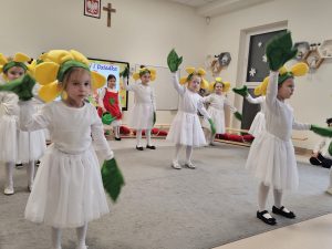 Dziewczynki ubrane na biało, na głowach kwiatki i liście na rękach tańczą na szarym dywanie.