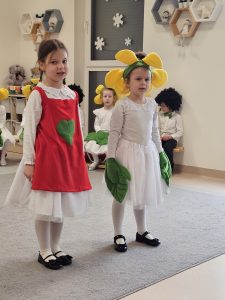 Dwie dziewczynki, jedna w czerwonym fartuszku, druga ubrana jak kwiatek recytują wiersz.