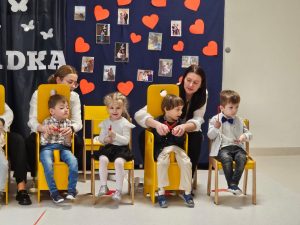 Dzieci siedzą na krzesełkach, w tle dekoracja i
