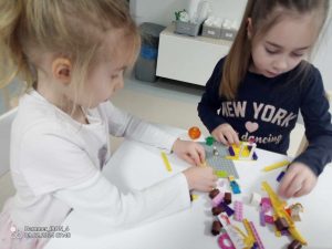 Dziewczynki przy stoliku tworzą budowle z klocków LEGO.
