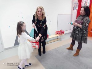 Pani dyrektor przedszkola wręcza dyplom i nagrodę dziewczynce ubranej na biało.