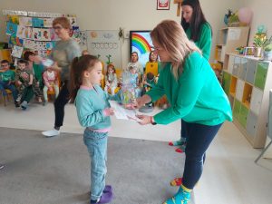 Pani dyrektor Izabela ubrana w zielony sweter i kolorowe skarpetki wręcza dziewczynce dyplom i nagrodę.