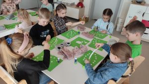 Dzieci siedzą przy stolikach i wykonują pracę plastyczną - zielony znaczek produktów ekologicznych.