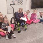 Dzieci siedzą na szarym dywanie w kole. Wśród dzieci siedzą dzieci niepełnosprawne np. na wózku.