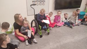 Dzieci siedzą na szarym dywanie w kole. Wśród dzieci siedzą dzieci niepełnosprawne np. na wózku.