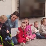 Dzieci siedzą na szarym dywanie w kole. Wśród dzieci siedzą dzieci niepełnosprawne np. na wózku.