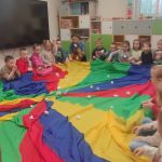 Dzieci siedzą w kole i trzymają kolorową chustę, którą poruszają do góry i na dół.