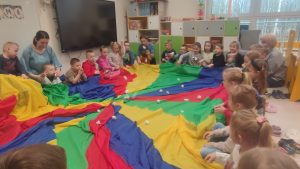 Dzieci siedzą w kole i trzymają kolorową chustę, którą poruszają do góry i na dół.