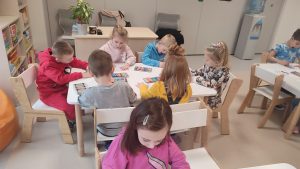 Dzieci siedzą przy białych stolikach i kolorują kolorowankę kredkami.