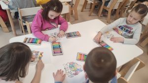 Dzieci siedzą przy białych stolikach i kolorują kolorowankę kredkami.