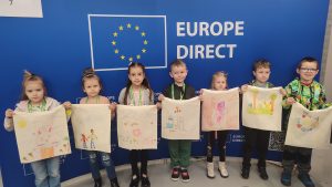 Dzieci na tle flagi Unii Europejskiej pozują do zdjęć z samodzielnie wykonaną torbą ekologiczną.