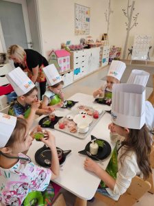 Dzieci w czapkach kucharskich siedzą przy białych stolikach i tworzą kolorowe desery z produktów ekologicznych.