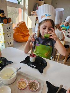 Dziewczynka próbuje zielony koktajl. Na stole leży talerzyk w kształcie kotka i smaczny deser.