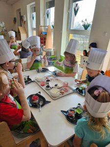 Dzieci w czapkach kucharskich siedzą przy białych stolikach i tworzą kolorowe desery z produktów ekologicznych.