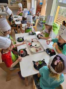 Dzieci w czapkach kucharskich siedzą przy białych stolikach i tworzą kolorowe desery z produktów ekologicznych.
