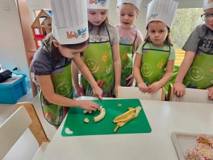Dziewczynka w czapce kucharskiej kroi nożem banana. Dziewczynki obserwują czynność.