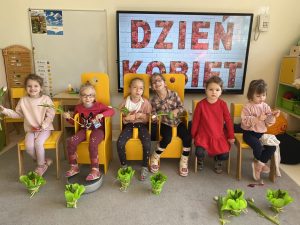 Sześć uśmiechniętych dziewczynek siedzi na krzesełkach. przed nimi stoją upominki- kwiatki hiacynty.