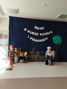Dziewczynka przebrana za kurkę zamiata podłogę, niedaleko stoi dziewczynka przebrana za zajączka podtrzymywana przez nauczyciela.
