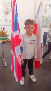 Chłopiec w białej koszuli stoi pomiędzy flagą Polski i Wielkiej Brytanii.