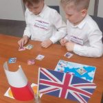 Dwóch chłopców w białych koszulach przy stole z flagą Wielkiej Brytanii rozwiązuje zadania.