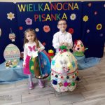 Chłopiec i dziewczynka pozują z dużymi pisankami na tle granatowej dekoracji.