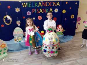 Chłopiec i dziewczynka pozują z dużymi pisankami na tle granatowej dekoracji.