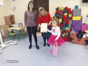 Dziewczynka w różowej spódnicy trzyma dyplom w ręce i stoi wśród dwóch pań.