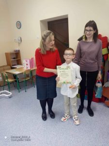 Chłopiec w białej koszuli odbiera dyplom z rąk dwóch pań.