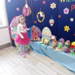 Dziewczynka na tle niebieskiej dekoracji i konkursowych pisanek pozuje do zdjęcia.