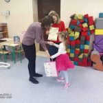 Dziewczynka w różowej spódnicy odbiera nagrodę i dyplom z rąk dwóch kobiet.