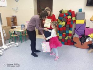 Dziewczynka w różowej spódnicy odbiera nagrodę i dyplom z rąk dwóch kobiet.