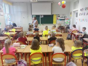 Dzieci w żółtych kamizelkach z przedszkola i uczniowie siedzą w ławkach szkolnych. Słuchają nauczyciela.