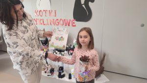 Dziewczynka otrzymuje gratulacje, dyplom , roślinkę w doniczce i kostkę Rubika z rąk nauczycielki.