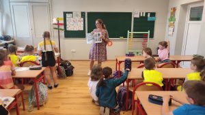 Pani nauczycielka stoi przed klasą dzieci i tłumaczy lekcję.