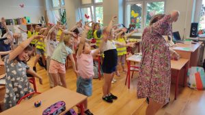 Wszystkie dzieci stoją między ławkami i naśladują ruchy nauczyciela.