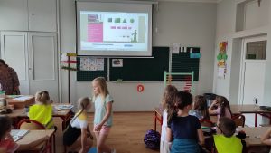 Dzieci siedzą w ławkach i patrzą na tablice multimedialną z dodawaniem.