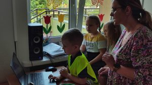 Dziecko w żółtej kamizelce pracuje w komputerze. Obok stoją dwie dziewczynki ze szkoły i nauczyciel.