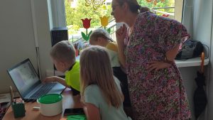 Dziecko w żółtej kamizelce pracuje w komputerze. Obok stoją dwie dziewczynki ze szkoły i nauczyciel.